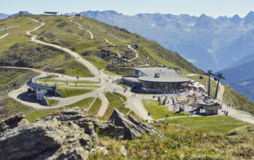 Panoramabild einer Bergstation der Fisser Bergbahnen. Sonnenschein, Bergpanorama und viele Besucher.