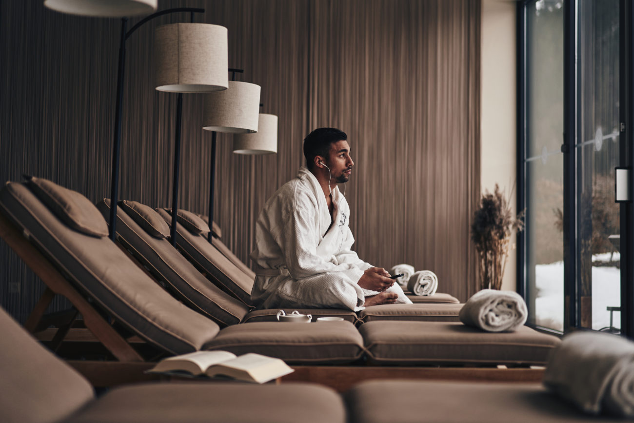 Ein Mann im Ifen Hotel Kleinwalsertal, der im Wellnessbereich auf einer Liege sitzt und Musik hört im Bademantel