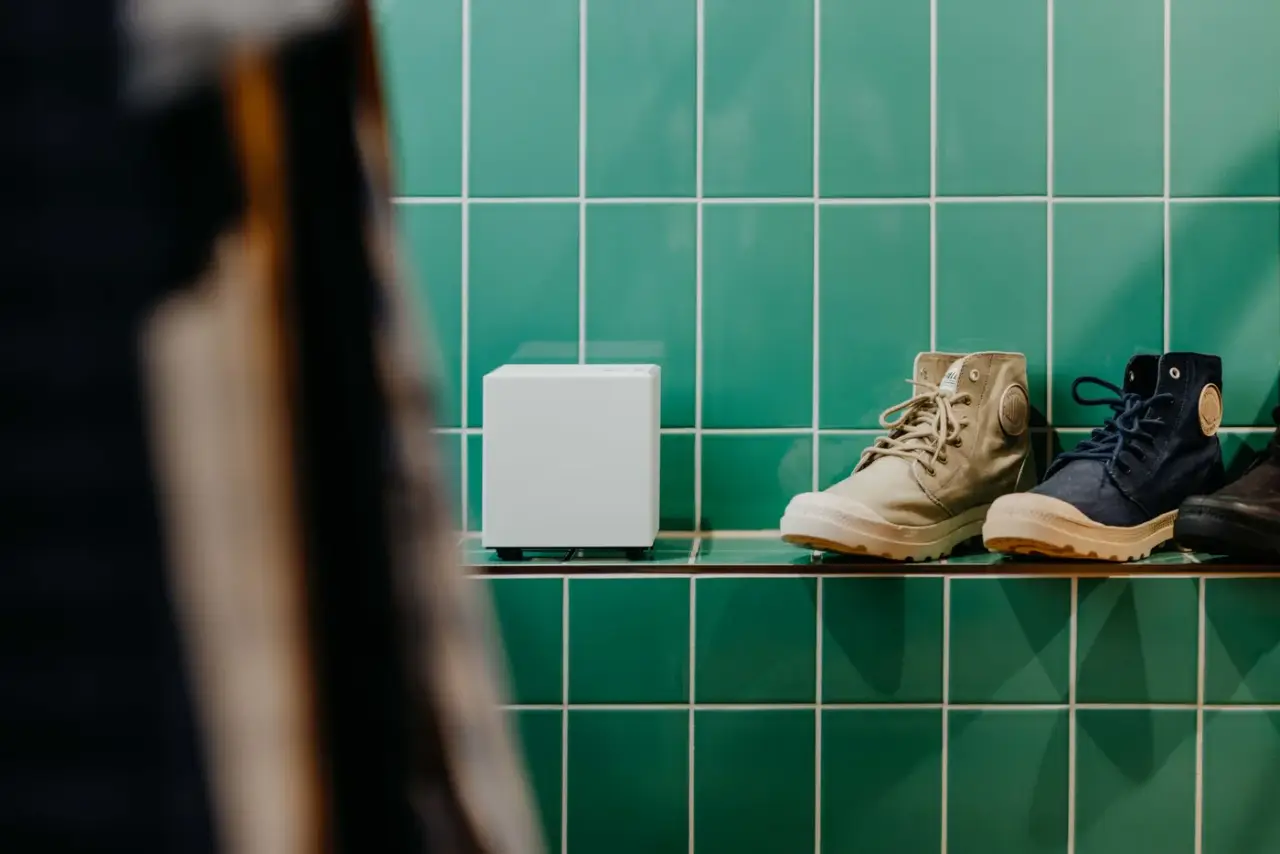 Weißes AROMEA Duftgerät neben Schuhen auf einem Regal vor grünen Fliesen in einem Showroom