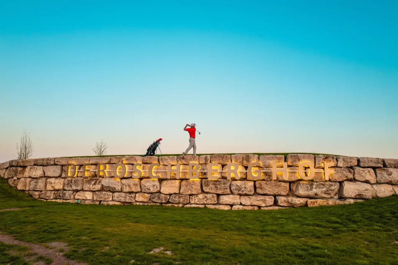 Golfer auf der Natursteinmauer mit dem Schriftzug des Öschberghofs im Abendlicht
