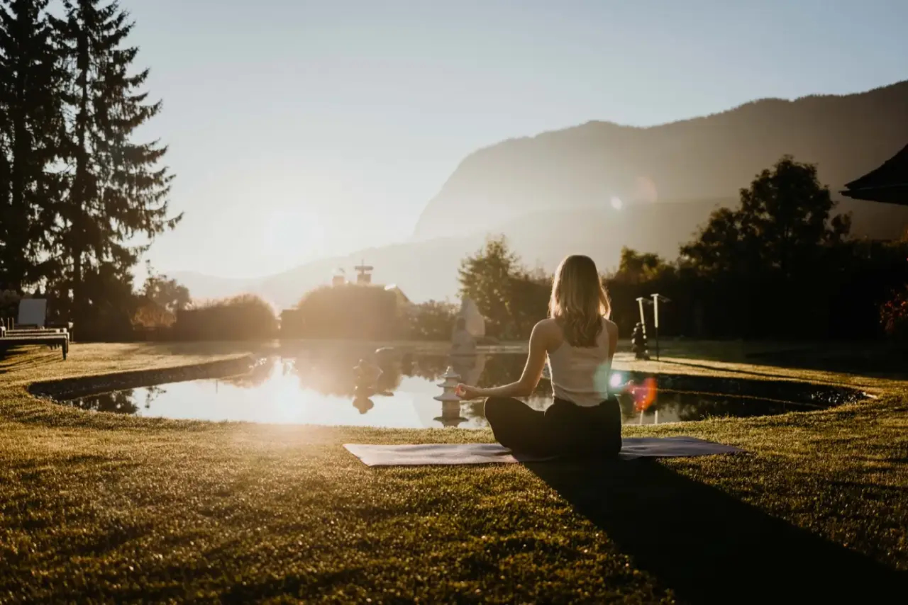 Frau sitzt im Gegenlicht im Lotussitz auf einer Yogamatte an einem Teich, im Hintergrund Berge
